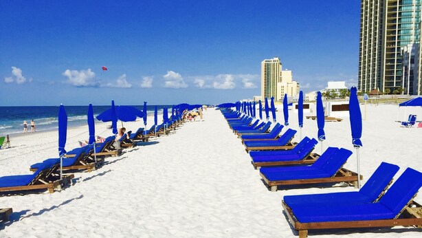 Uma praia com fileiras de espreguiçadeiras e guarda-sóis azuis, céu claro e um prédio alto de hotel ao fundo.