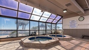 Uma área de spa com duas banheiras de hidromassagem, uma janela grande com vista para palmeiras e um gazebo, e um relógio na parede.