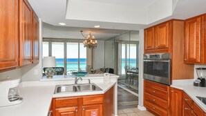 A kitchen with wooden cabinets, a stainless steel sink, and a coffee maker. There is a view of the ocean through large windows. - Beachfront elegance w/3 beds, 3 baths, & private balcony! (Destin)