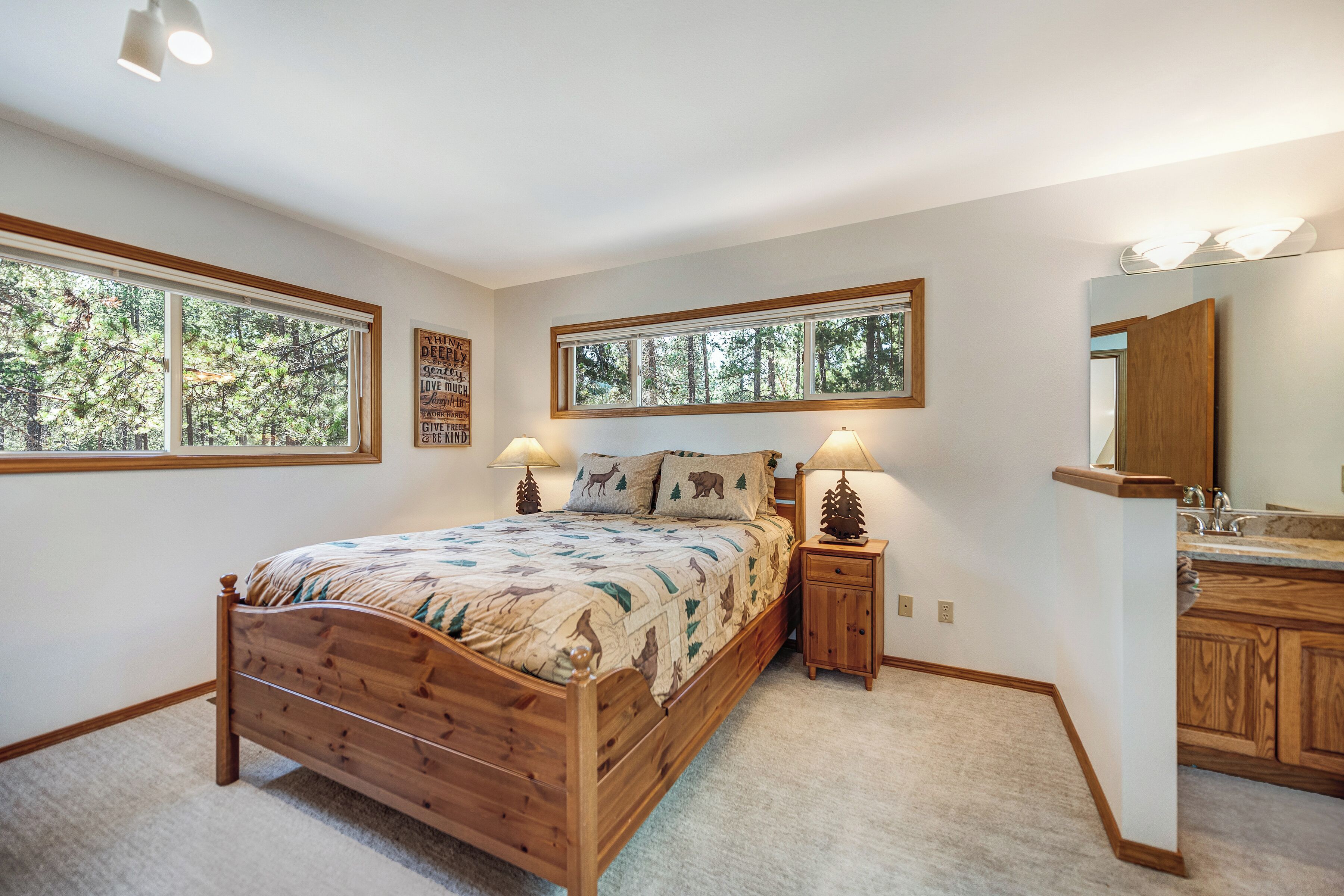 Une chambre à coucher avec un lit en bois, deux tables de chevet avec des lampes, une commode, un grand miroir et une fenêtre donnant sur des arbres.