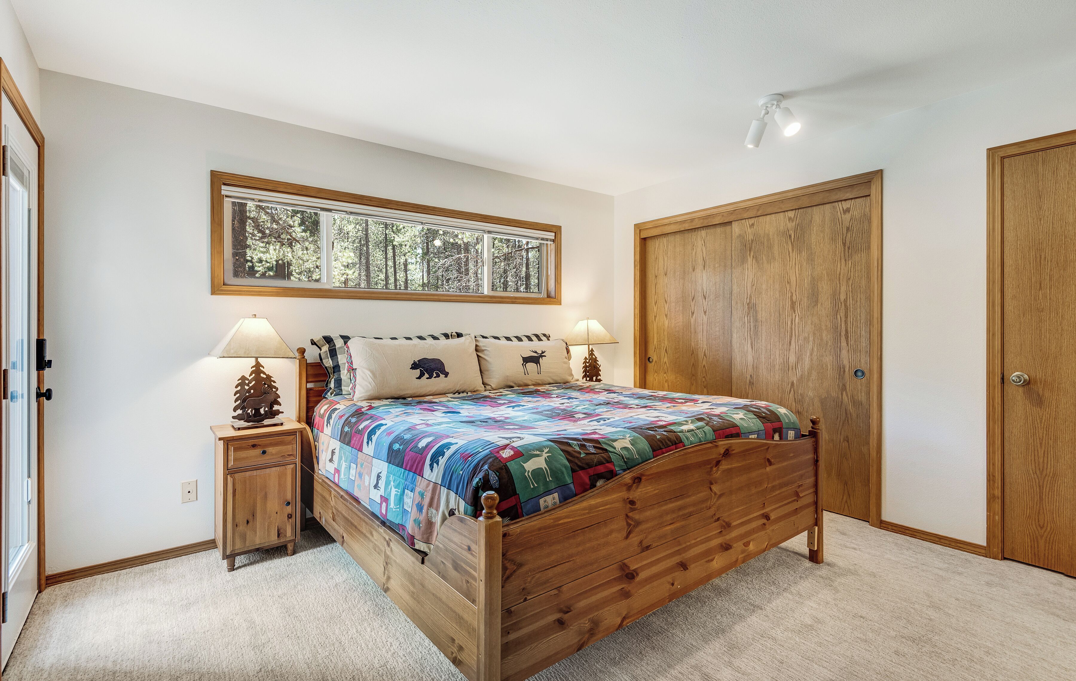 A bedroom with a wooden bed, a nightstand with a lamp, a window with a view of trees, and a wooden wardrobe.