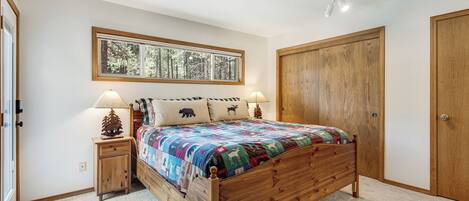 A bedroom with a wooden bed, a nightstand with a lamp, a window with a view of trees, and a wooden wardrobe.