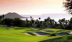 Ein Golfplatz mit Sandbunkern, Palmen und Blick auf den Ozean.