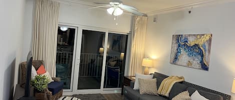 Ein Wohnzimmer mit grauem Sofa, einem Korbstuhl, einem kleinen Hocker und einem Deckenventilator.