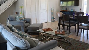 A living room with a sofa, ottoman, coffee table, and a dining area with chairs and a table. There is a TV mounted on the wall and a door with a shutter. - Oceanside, pet friendly, 5/3 smart home with paddle equip.-SUPs, kayaks & canoe (SugarLoaf)