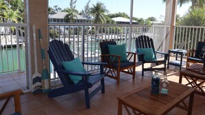 A covered outdoor seating area with two wooden chairs and a table, overlooking a pool and palm trees. - Oceanside, pet friendly, 5/3 smart home with paddle equip.-SUPs, kayaks & canoe (SugarLoaf)
