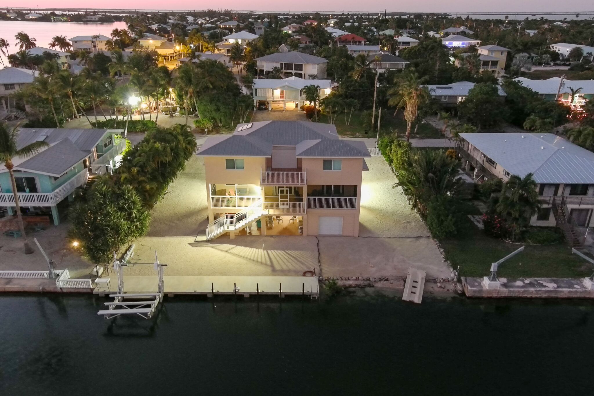 A waterfront property with a large house featuring a balcony, a dock, and a well-lit driveway.
