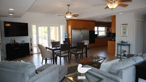 A spacious living area with a sofa set, a coffee table, and a television mounted on the wall. There is a dining area with chairs and a table, and a kitchen with cabinets and a refrigerator. - Oceanside, pet friendly, 5/3 smart home with paddle equip.-SUPs, kayaks & canoe (SugarLoaf)