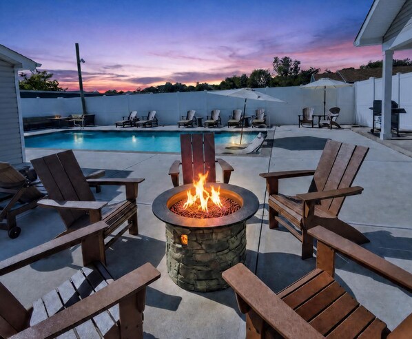 An outdoor pool area with a stone fire pit surrounded by wooden Adirondack chairs.