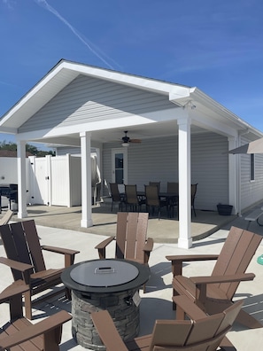 A covered patio area with Adirondack chairs, a stone fire pit, and a dining table with chairs.