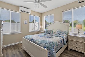 A bedroom with a bed, a nightstand, a lamp, a ceiling fan, and large windows with blinds.