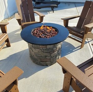 A circular stone fire pit with a black top surrounded by wooden Adirondack chairs.