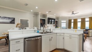 Une cuisine moderne avec des meubles blancs, un lave-vaisselle et un grand îlot central doté d’un évier. On y trouve également un ventilateur de plafond, un éclairage encastré et une vue sur l’océan grâce à de grandes fenêtres.