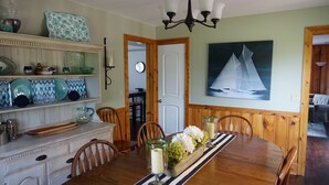 Un coin repas avec une table en bois, des chaises, un lustre et un tableau représentant des voiliers accroché au mur.