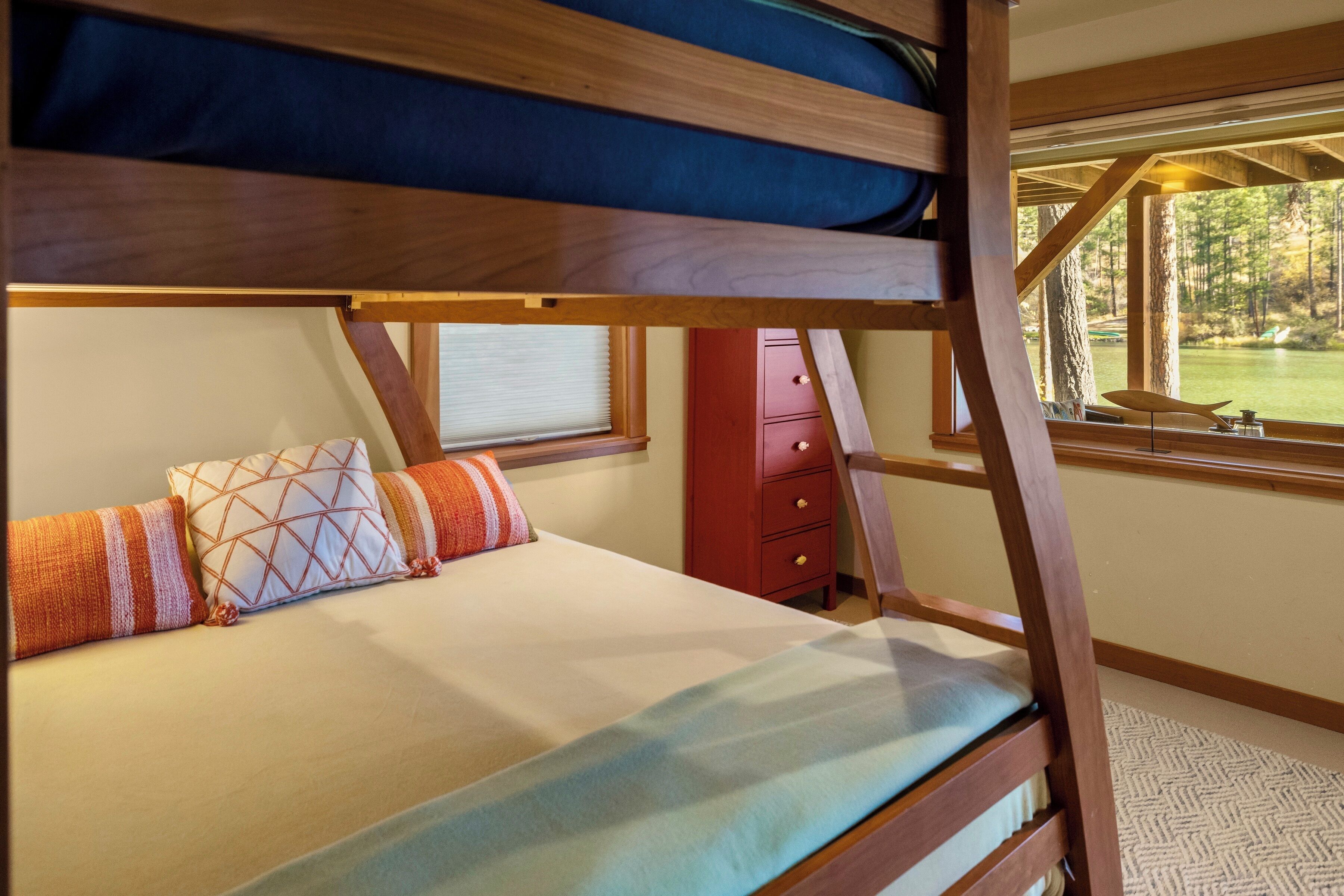 Una litera con estructura de madera, una cama con colchón color beige y almohadas estampadas, un aparador rojo y una ventana con vistas a árboles y un lago.