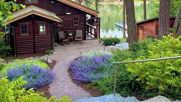 Une cabane dotée d’une véranda et d’un espace salon, entourée d’une végétation luxuriante et bordée d’un lac.