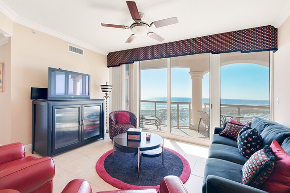 A spacious living room with a large window offering a view of the ocean, a ceiling fan, a television, and a dining area with a round table. - Gulf front, Portofino paradise, sunset balcony (Pensacola Beach)
