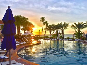 A resort pool with lounge chairs and umbrellas, surrounded by palm trees and a sunset view. - Gulf front, Portofino paradise, sunset balcony (Pensacola Beach)