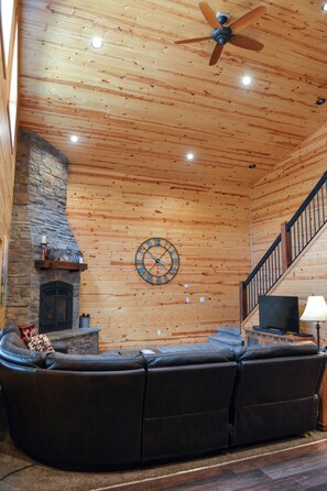 A wooden interior with a stone fireplace, a curved leather sofa, a ceiling fan, and a staircase.