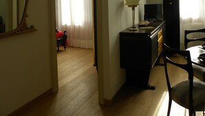 Une pièce avec un plancher en bois, un bureau avec une lampe, une télévision et une chaise. Il y a un miroir au mur et une fenêtre avec des rideaux.