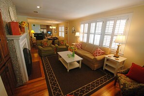 Cozy living room with a fireplace, comfortable seating, and a TV, featuring a large window with white shutters.