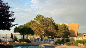 Una piscina con borde de ladrillo, sillas de descanso y un área cercada con árboles.