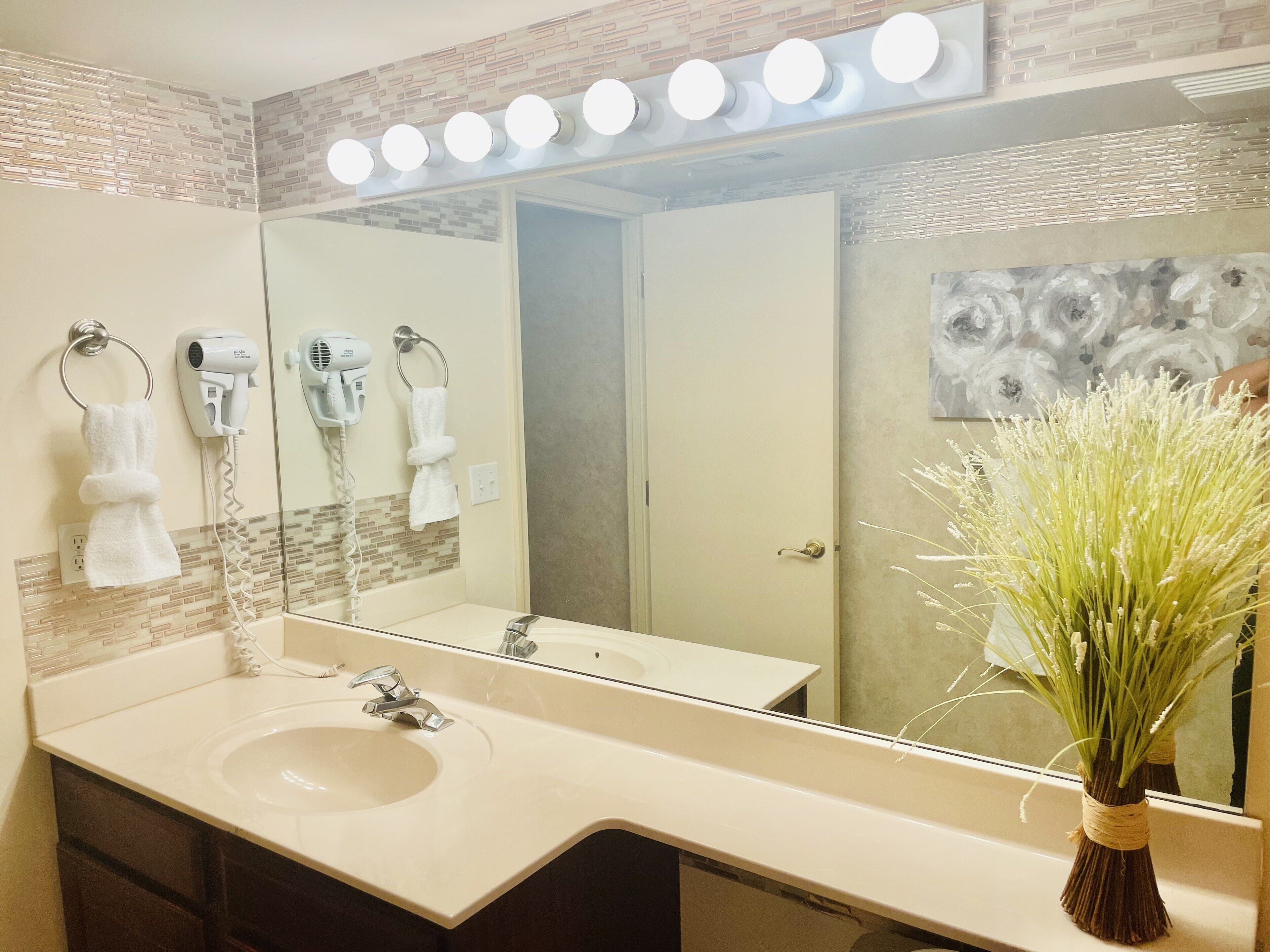 Un baño con un espejo grande, un lavabo, dos secadores de pelo y un jarrón con flores.