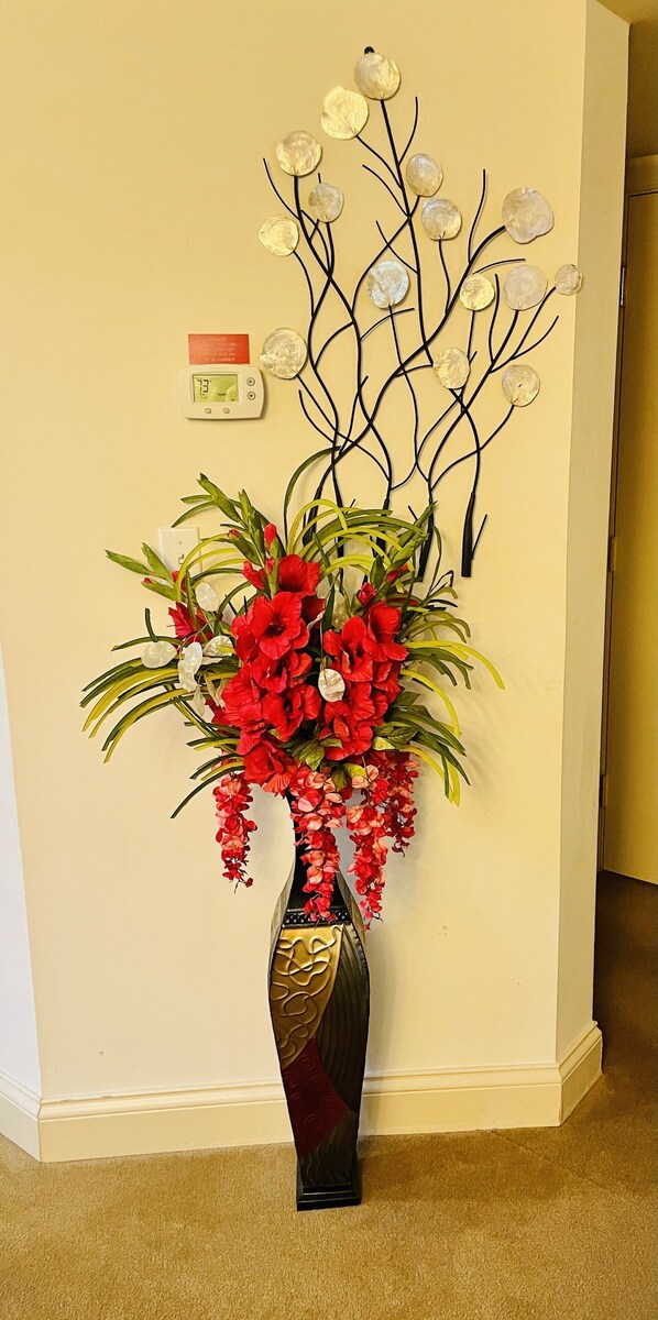 Una esquina de pared con una escultura decorativa de metal con forma de árbol, un termostato y un jarrón con flores rojas.
