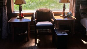 Ein gemütliches Zimmer mit einem großen Fenster und Blick auf eine grüne Landschaft. In der Mitte befinden sich zwei Holztische mit Lampen und ein Stuhl.