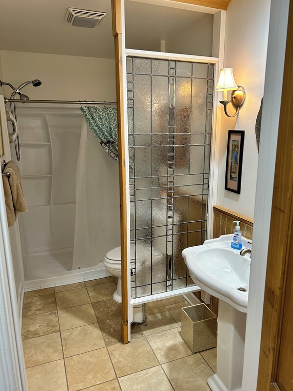 Une salle de bain avec une douche à parois de verre, des toilettes, un lavabo avec un distributeur de savon et un luminaire fixé au mur.