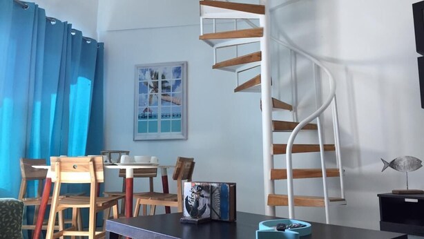 A room with a spiral staircase, wooden chairs, a table with a book, and a framed picture on the wall.