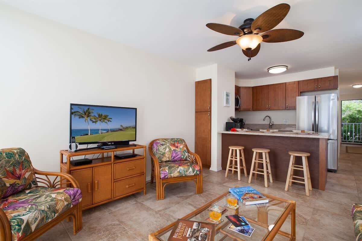 Una sala de estar con televisión, dos sillas de mimbre, una mesa de centro con libros y vasos, un ventilador de techo y una cocina con refrigerador y taburetes de bar.
