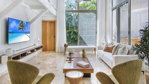 Ein geräumiges Wohnzimmer mit großem Fernseher, Deckenventilator, weißem Sofa und einem Essbereich mit Holztisch.