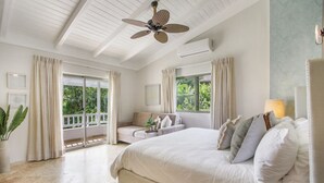 Ein Schlafzimmer mit einem großen Bett, einem Deckenventilator, einem Balkon mit Vorhängen und Blick ins Grüne.
