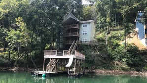 A multi-level wooden structure with a deck and stairs, surrounded by dense forest and a body of water. - Super Location in Lakeside Estates--w/pool; basketball ct; walking tr; boat ramp (Lafollette)