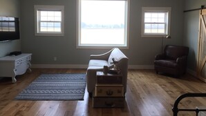 A spacious living room with a large window, a flat-screen TV, a leather armchair, and a wooden floor. - Relax At The Farm!   Newly Built Studio Apartment In The Barn At Selah Farm (Kalispell)
