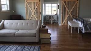 A spacious room with a large sliding barn door, a cozy sofa, a desk with a chair, and a bed with a metal frame. - Relax At The Farm!   Newly Built Studio Apartment In The Barn At Selah Farm (Kalispell)