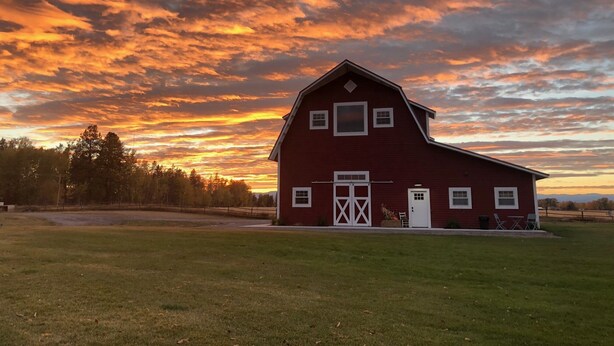 A red barn with a white door and windows, surrounded by a grassy field and trees, under a sunset sky. - Relax At The Farm!   Newly Built Studio Apartment In The Barn At Selah Farm (Kalispell)