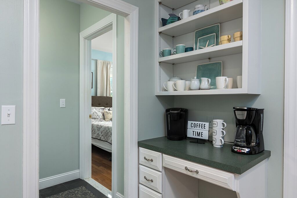 A kitchen with a coffee station, a bed visible through an open door, and a shelf with kitchenware.