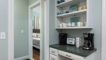 A kitchen with a coffee station, a bed visible through an open door, and a shelf with kitchenware.