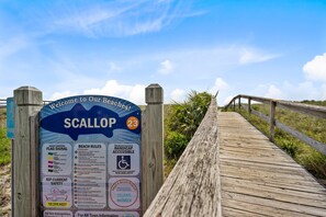 A wooden boardwalk leading to a beach entrance sign with information on beach conditions, rules, and safety.