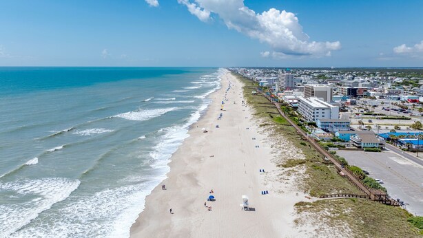 A beach with a long stretch of sand, a few umbrellas, and buildings along the shore. - "Sand Dollar Suites" suite 1: A short stroll from our sandy shores! (Carolina Beach)