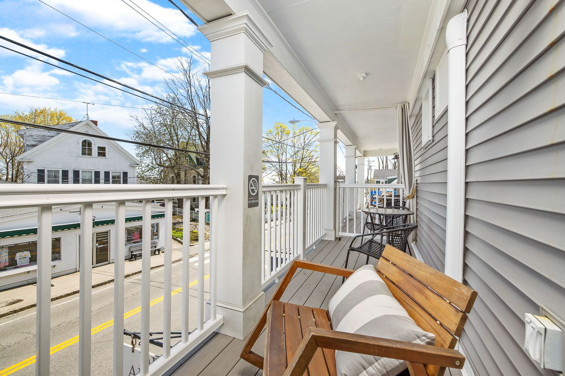 Eine hölzerne Bank auf einer Veranda mit weißer Holzbalustrade, mit Blick auf eine Straße, auf der sich ein Zug und ein Gebäude befinden.