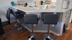 Cocina moderna con una isla central, dos taburetes de bar, una mesa de comedor y un pequeño rincón de estar.