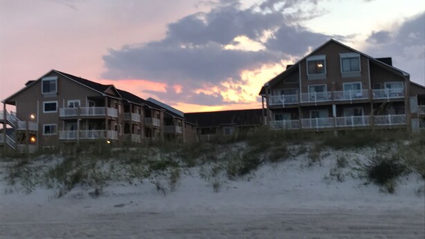 Un hotel frente a la playa con varios pisos y balcones, ubicado en terreno arenoso con dunas al frente.