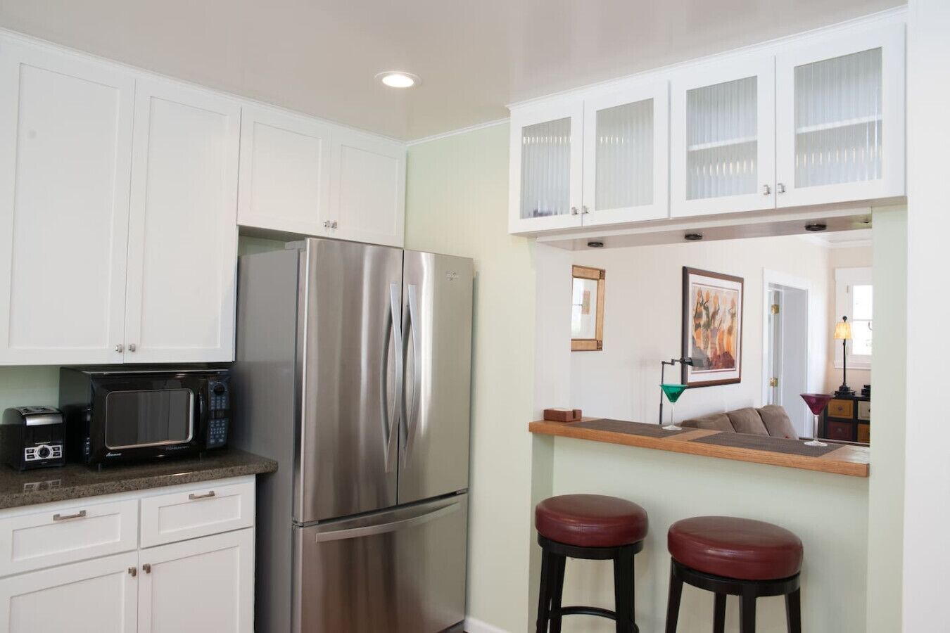 A compact kitchen with white cabinets, a stainless steel refrigerator, a microwave, and two stools.