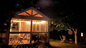 Une petite cabane en bois, illuminée de l’intérieur et dotée d’une façade en verre, entourée d’arbres et baignée par la clarté d’un ciel nocturne.