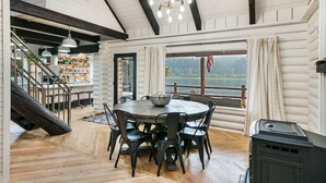 A spacious dining area with a round table and chairs, a view of a lake, and a rustic interior with exposed wooden beams and a fireplace.