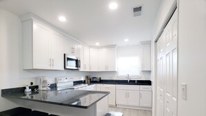 A modern kitchen with white cabinets, a black countertop, and a stainless steel range hood. There are three bar stools and a trash can. - Beautiful Oceanside Property W/Pool in Great Beach Location! (Corolla)