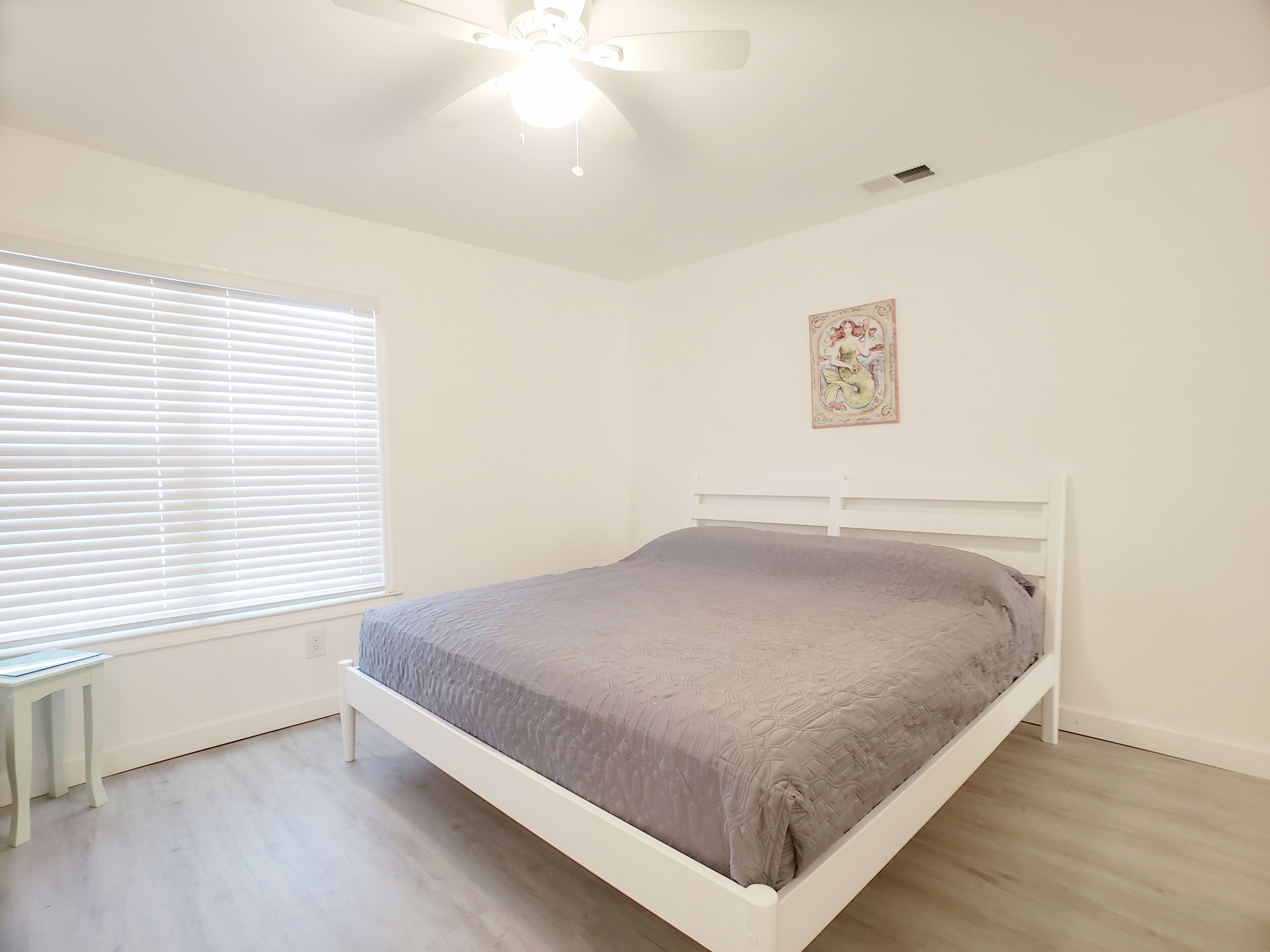 Un dormitorio con cama, una ventana con persianas, un ventilador de techo y un cuadro colgado en la pared.